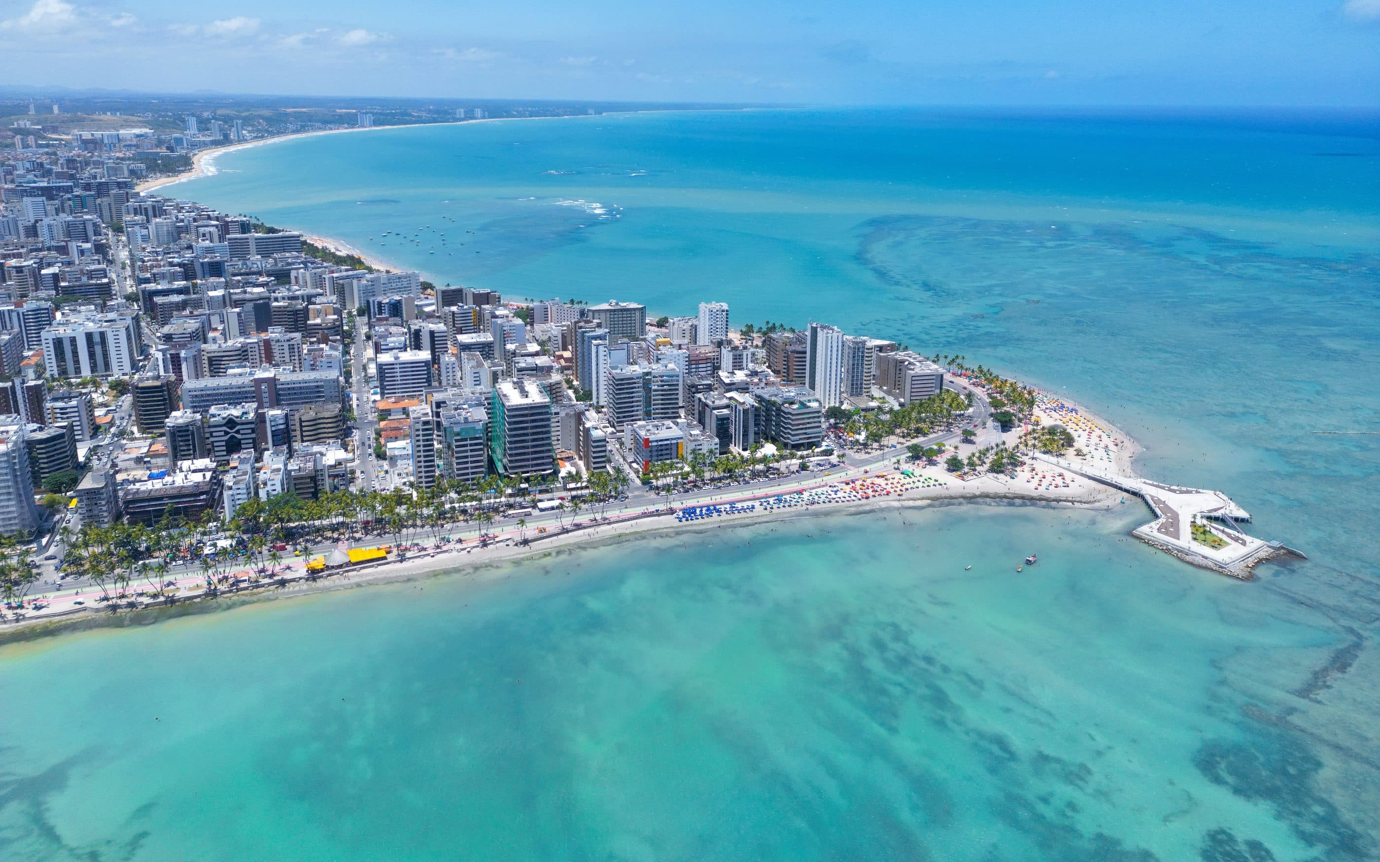 Vista aérea da orla de Maceió: os prédios da praia de um lado e o mar esverdeado sobre os recifes do outro